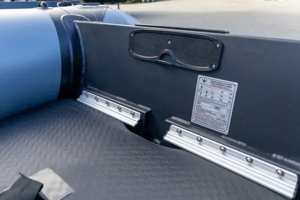 Close-up of the rear deck of a black inflatable boat, showing the textured non-slip mat, silver metal rails, a black speaker grille, and a product information sticker.