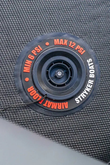 A close-up of a circular black air mat valve assembly with red and white text reading 'MIN 6 PSI', 'MAX 12 PSI', 'AIRMAT FLOOR', and 'STRYKER BOATS', set against a dark textured mat surface.